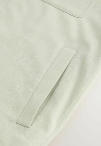 Tela texturizada de color verde claro con un acabado elegante. Presenta una solapa de bolsillo rectangular integrada en el diseño.