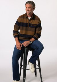Pullover mit Viertelreißverschluss in Braun- und Navystreifen, strukturiertem Stoff, Brusttasche, kombiniert mit blauen Jeans und weißen Sneakers, auf einem Hocker sitzend.