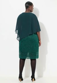 Zelené krajkové šaty s chiffonovým překryvem. Mají přiléhavý krajkový design a zvlněný lem. Nosí se s černými punčochami a podpatky.
