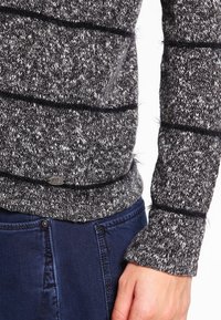 Sweter dzianinowy w ciemnoszarym kolorze z jasnoszarymi plamkami. Posiada teksturowane paski oraz małą metalową metkę z logo u dołu.