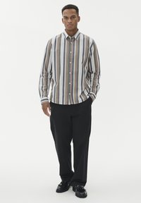 Moški stoji v črnih hlačah, črnih čevljih s peresi ter bež, beli, sivi in mornarsko modri majici s verticalnimi črtami na gumb.