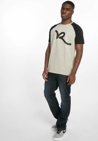 Grå bomull T-skjorte med svarte raglanermer og et stort svart grafikk på forsiden, kombinert med mørke jeans og grå joggesko.