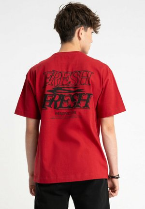 Młody mężczyzna stojący tyłem, ubrany w czerwoną koszulkę z dużym czarnym napisem „FRESH” i abstrakcyjnym wzorem na plecach, w połączeniu z czarnymi spodniami.
