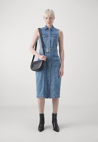 Abito in denim con design senza maniche, apertura frontale con bottoni, due tasche sul petto e vita con cintura. Abbinato a una borsa a tracolla nera e stivali alla caviglia.