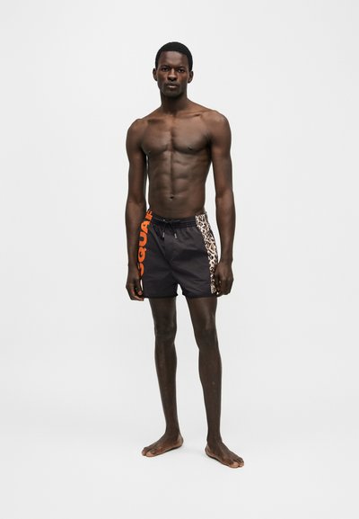 Άντρας χωρίς πουκάμισο, όρθιος με γυμνά πόδια, φοράει μαύρα μαγιό με πορτοκαλί κείμενο και πάνελ πλευρών με τύπωμα λεοπάρδαλης σε απλό λευκό φόντο.