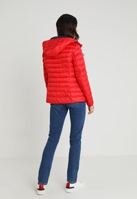 Vrouw in een rode gewatteerde jas met een capuchon, blauwe jeans en witte sneakers, staand met haar rug naar de camera tegen een effen achtergrond.