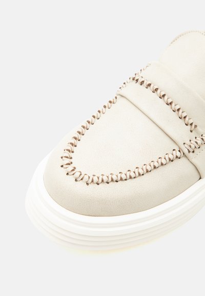 Μπεζ παπούτσι τύπου loafer με διακοσμητική ραφή γύρω από τη μύτη και παχιά λευκή σόλα με ραβδώσεις, εμφανίζεται σε κοντινό πλάνο.