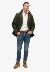 Abrigo de marina verde oliva sobre una camisa blanca, combinado con jeans azules y botas beige. Presenta botones de doble botonadura y un cinturón marrón.