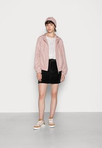 Casaco corta-vento rosa claro com capuz, punhos elásticos, acompanhado por uma t-shirt branca cropped e uma saia preta, complementado por sapatilhas beijas e brancas.