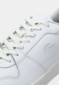White leather sneakers with textured surface, flat white laces, a subtle embossed logo, and stitched detailing on the sides.
