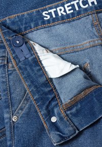 Jean en denim bleu partiellement déboutonné montrant la doublure blanche de la poche avec un bouton et le mot « STRETCH » imprimé sur la ceinture.