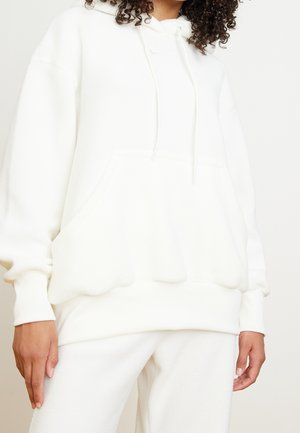 Person iført oversized hvid hættetrøje og matchende hvide bukser, der viser afslappet hverdagsoutfit fra midt på låret til halsen.