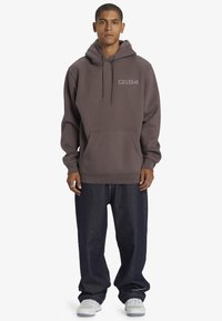Grijze hoodie met koordjes en een voorzak, gecombineerd met donkere denim jeans. Heeft een logo in een lichter tint op de borst.