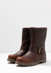 Paire de bottines en cuir marron foncé avec bride et boucle, bouts ronds et semelles crantées en caoutchouc sur fond blanc uni.
