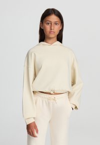 Felpa corta color crema con cordino in vita, maniche lunghe e cappuccio. Abbinata a pantaloni della tuta con cordino coordinati.