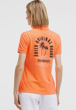 Frau mit hellbraunen Haaren trägt ein orangefarbenes Poloshirt mit Palmenmotiv und Text auf dem Rücken, kombiniert mit weißen Shorts.