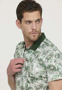 Polo à manches courtes avec un motif de feuillage vert sur fond blanc, doté d'un col vert et d'une patte de boutonnage à trois boutons.