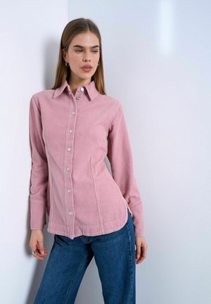 Kvinde, der læner sig op ad en væg iført en tætsiddende pink fløjls-skjorte med knapper og blå jeans, der kigger en smule til siden.