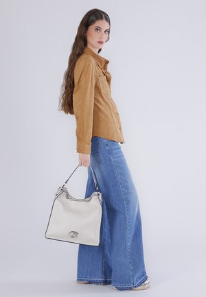 Donna con capelli lunghi che indossa una camicia color cammello abbottonata e jeans blu a gamba larga, con una grande borsa bianca tenuta di lato.