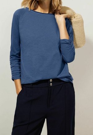 Vrouw draagt een blauw shirt met lange mouwen en een zwarte broek, houdt een beige gestructureerd jasje over haar schouder.
