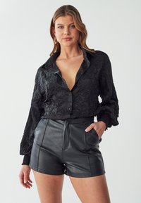 Zwarte, gestructureerde button-up shirt met een diepe V-hals en lange mouwen, gecombineerd met high-waisted zwarte leren short met voornaden.