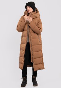 Lange bruine pufferjas met een capuchon, met horizontaal gestikte delen, ritsen en een relaxte pasvorm. Draag het over een zwarte broek en laarzen.
