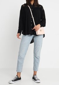 Una donna sta in piedi con sicurezza, indossando una camicia nera a bottoni, jeans azzurri chiari e sneaker nere, tenendo una piccola borsa a tracolla rosa.