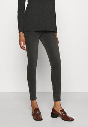 Woman wearing fitted dark gray jeans, black long-sleeve top, and shiny brown heeled loafers, standing on light gray floor against white wall.