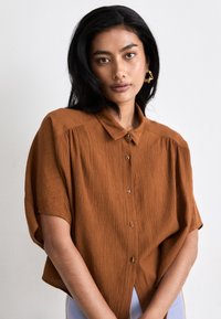 Brown short-sleeve button-up shirt with a textured fabric, featuring a relaxed fit and classic collar. Decorative buttons enhance design.