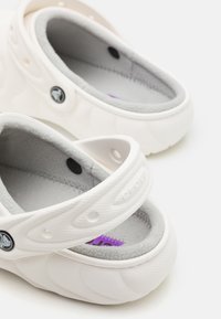 Chaussures blanches avec un dessus texturé, intérieur doux gris, bande ajustable et détails d'accent violet ; design léger et antidérapant.