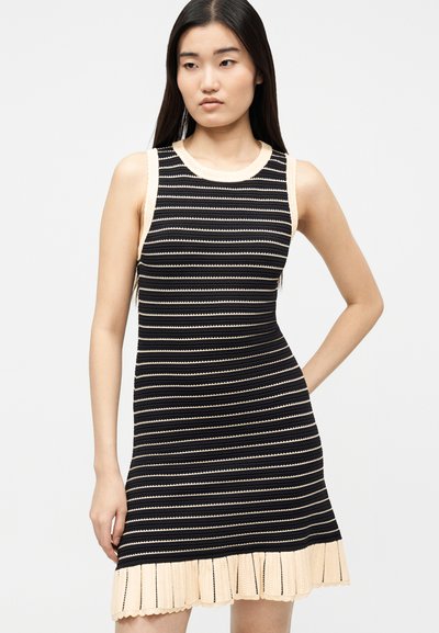 Νεαρή γυναίκα με μακριά σκούρα μαλλιά, που φοράει ένα αμάνικο μαύρο και μπεζ ριγέ φόρεμα με μπεζ βολάν στο τελείωμα.