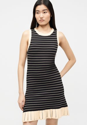 Joven con cabello oscuro y largo que lleva un vestido sin mangas de rayas negras y beige con un dobladillo beis con volantes.