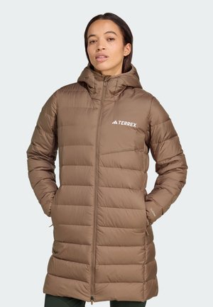 Mujer con una chaqueta acolchada marrón de la marca Terrex con capucha, manos en los bolsillos, de pie frente a un fondo claro.
