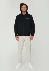 Svart zip-up jacka med hög krage och elastiska muddarna, med två fickor med dragkedja och en logotyp på bröstet. Bärs med ljusbeiga byxor.