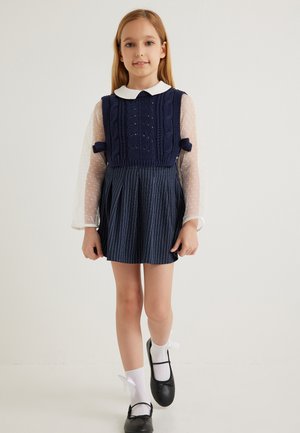 Vestido de punto azul marino con parte superior texturizada, cuello blanco y falda plisada a rayas. Lleva puesto calcetas blancas y zapatos negros.