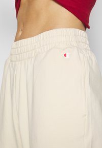 Pantalones de chándal color crema con una cinturilla elástica fruncida, que presenta un pequeño logo rojo y blanco en la cadera. Tejido suave y texturizado.