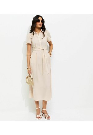 Kobieta w beżowej sukience midi z paskiem w talii, białych sandałkach na obcasie, okularach przeciwsłonecznych, złotych bransoletkach, trzymająca plecioną torebkę na tle białego tła.