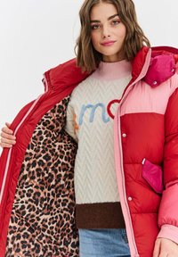 Žena v červené a růžové péřové bundě, která odhaluje leopardí vzor podšívky, s bílým svetrem se barevným vyšíváním a modrými džíny.