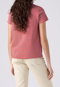 Personne aux longs cheveux bouclés portant une chemise à manches courtes rayée rouge et blanche et un pantalon beige clair, vue de dos.