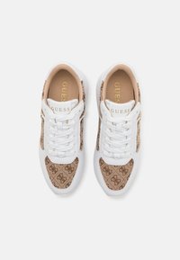 Baskets avec une base synthétique blanche, des panneaux latéraux en textile à motif marron, un laçage à l'avant et un logo "GUESS" doré sur la languette.