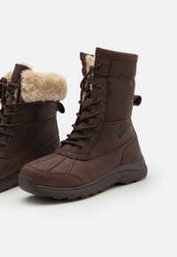 Bruine leren laarzen met nepbontkraag, vetersluiting, rubberen zool en gestikte accenten, met een waterdicht label aan de zijkant.