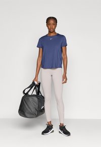 Μπλε ναυτικού χρώματος μπλουζάκι Nike, ανοιχτό μπεζ κολάν, μαύρα αθλητικά παπούτσια και μαύρη τσάντα duffel με λευκό λογότυπο. Απλό φόντο.