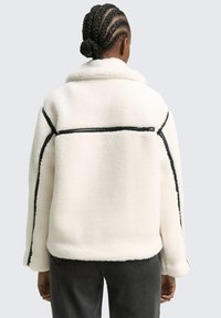 Jachetă albă din fleece, cu detalii din piping negru, design cu guler și o croială lejeră. Prezintă o suprafață texturată și mâneci supradimensionate.