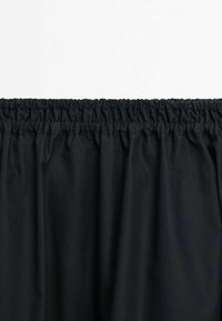 Tissu noir avec ceinture élastique froncée, créant des plis régulièrement espacés le long du bord supérieur.