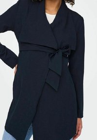 Donkerblauwe wikkeljas met een gestructureerde stof, voorzien van een strik aan de voorkant en lange mouwen, met een licht asymmetrische zoom.
