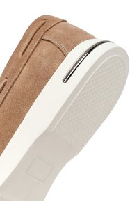 Zapatilla de ante color canela con suela de goma blanca texturizada y detalle metálico cerca del talón, mostrando un diseño superior con costuras.