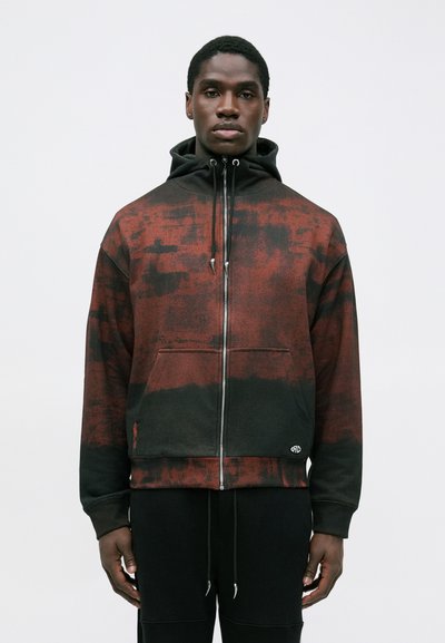 Mężczyzna w czerwono-czarnym rozpinanym hoodie z przednią kieszenią i czarnymi sznurkami, w zestawieniu z czarnymi spodniami, stojący na tle gładkiej ściany.