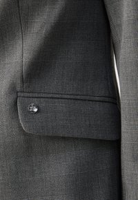 Gros plan d'une poche de blazer à carreaux gris avec un rabat et un petit bouton métallique rond.