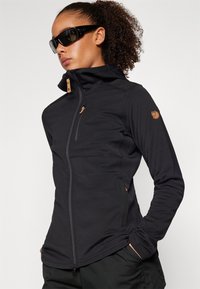 Giacca nera aderente con colletto alto, tasche con zip e dettagli in pelle marrone. Realizzata in un materiale liscio ed elastico.