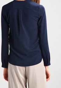 Blusa a maniche lunghe blu navy con scollatura rotonda, dettaglio plissettato sul retro e polsini con bottoni, abbinata a pantaloni chiari.